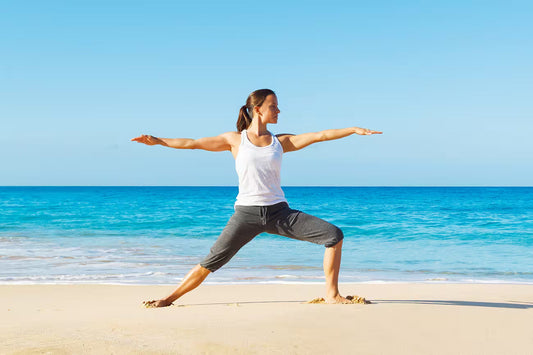 Person performing a warrior yoga pose on a sandy beach with arms extended, facing the ocean under a clear blue sky. Turquoise water and shoreline are visible in the background.